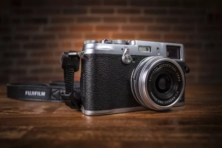 HD desktop wallpaper featuring a Fujifilm man-made camera with a silver and black body on a wooden surface against a blurred brick background.
