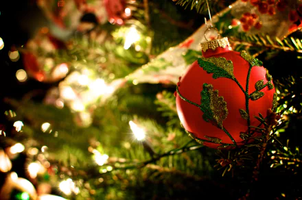 A vibrant red Christmas ornament with green detailing hangs from a lush tree, illuminated by warm lights, creating a festive holiday atmosphere in this HD desktop wallpaper.
