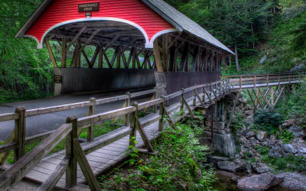 A vibrant covered bridge stands surrounded by lush greenery, with a wooden walkway leading across a serene stream, captured in stunning HD detail for a desktop wallpaper background.