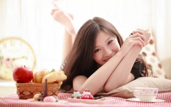 HD desktop wallpaper of a woman lying on a bed, smiling contentedly. A basket of breakfast items, including bread and an apple, is nearby, setting a cheerful, relaxed mood.