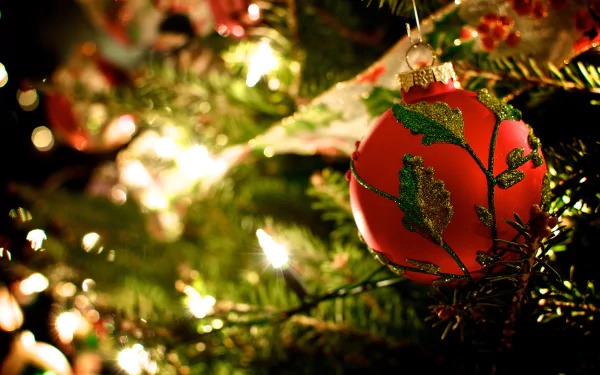 A vibrant red Christmas ornament with green detailing hangs from a lush tree, illuminated by warm lights, creating a festive holiday atmosphere in this HD desktop wallpaper.