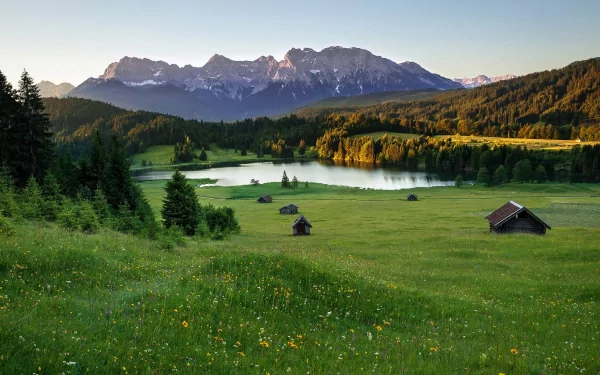 HD PC desktop wallpaper of a man-made cabin set in a lush green meadow with a lake and mountain range in the background under a clear sky.