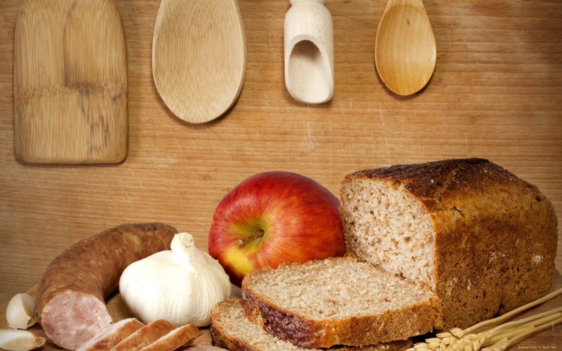 HD PC desktop wallpaper featuring a still life of sliced whole grain bread, an apple, garlic, sausage, and wooden kitchen utensils on a wooden surface.