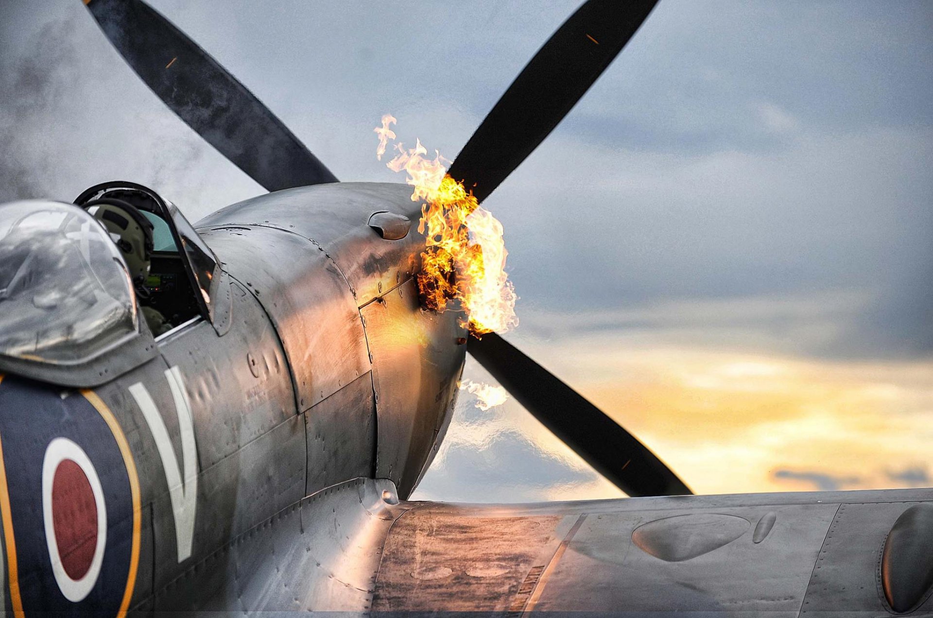 A dramatic close-up of a Supermarine Spitfire with flames erupting from its engine, set against a stunning sunset backdrop, serving as a striking HD desktop wallpaper.