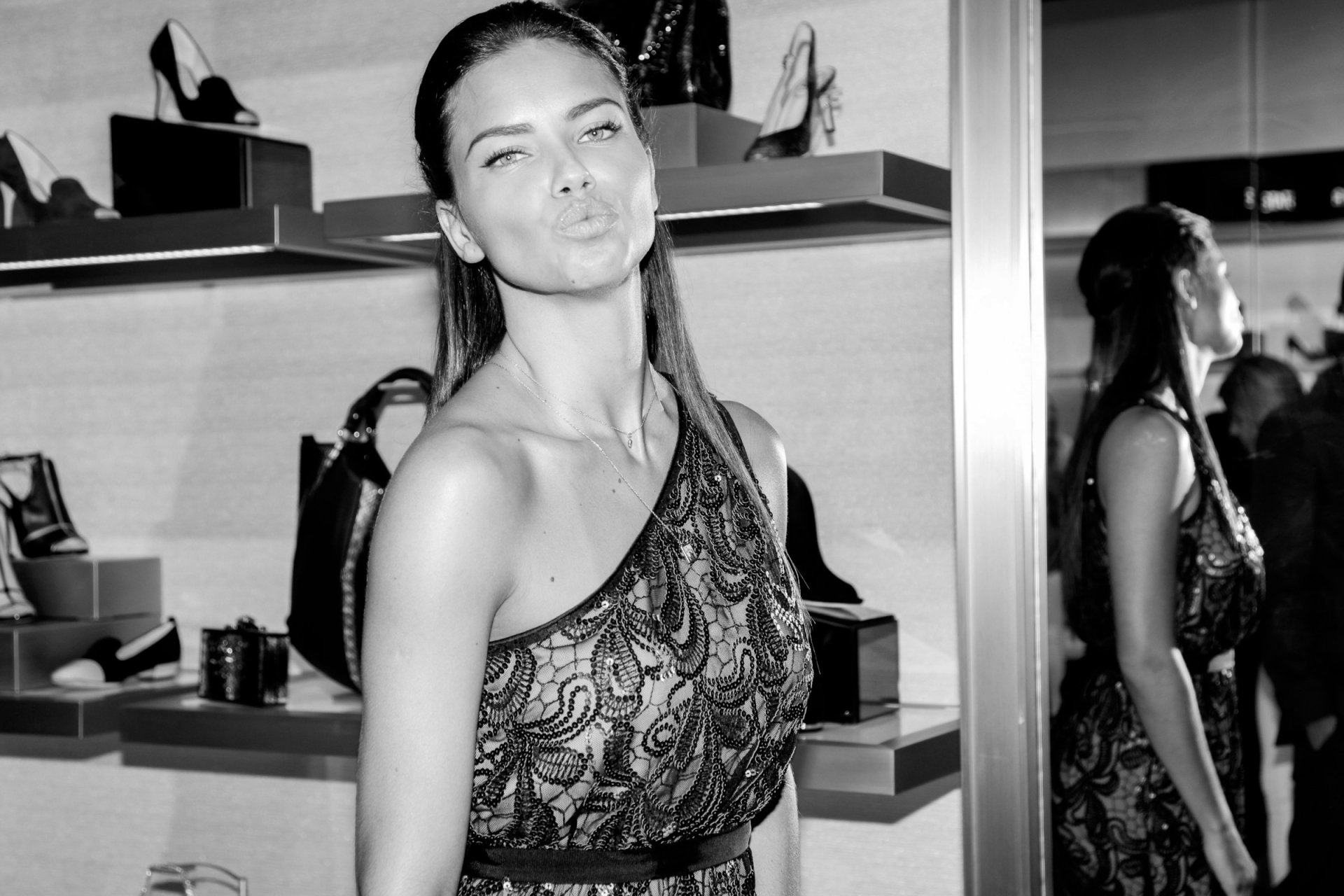 Black and white HD desktop wallpaper featuring celebrity Adriana Lima posing playfully in a stylish, patterned one-shoulder dress inside a chic boutique.