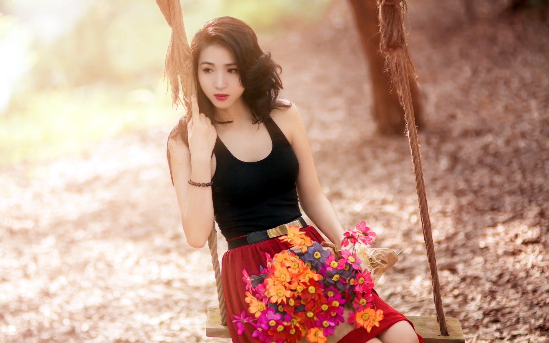 HD desktop wallpaper featuring an Asian woman in a black top and red skirt, holding flowers while sitting on a swing surrounded by a soft, sunlit background.