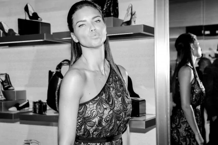 Black and white HD desktop wallpaper featuring celebrity Adriana Lima posing playfully in a stylish, patterned one-shoulder dress inside a chic boutique.