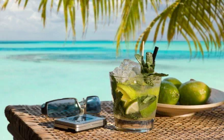 HD desktop wallpaper featuring a refreshing cocktail with lime and mint on a wicker table by the turquoise ocean, framed by palm leaves and accompanied by sunglasses and a phone.