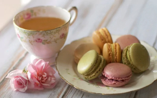 A charming arrangement of colorful macarons on a plate beside a delicate floral teacup and soft roses, creating a delightful food-themed HD desktop wallpaper.