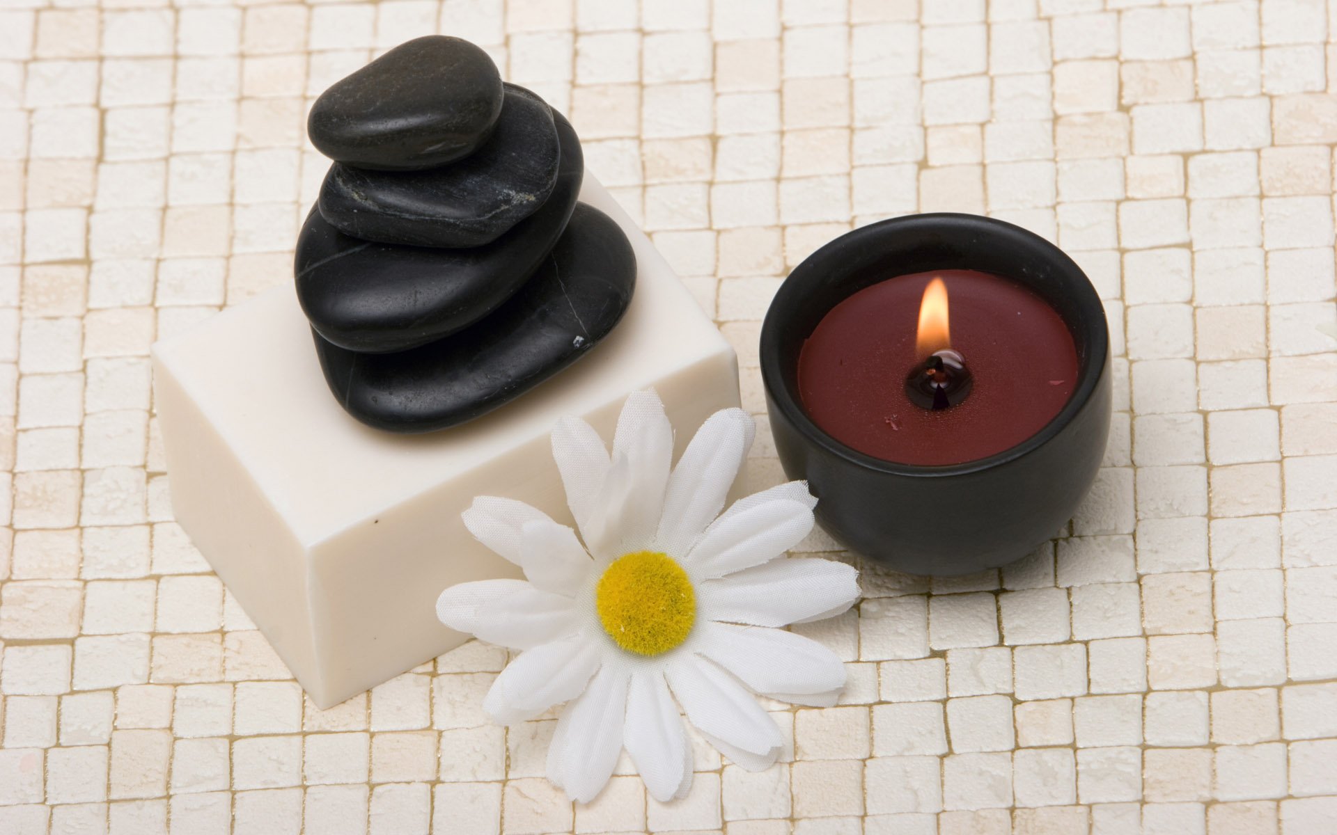 HD desktop wallpaper of a man-made spa scene featuring stacked black stones on soap, a white daisy, and a lit candle on a tiled surface.