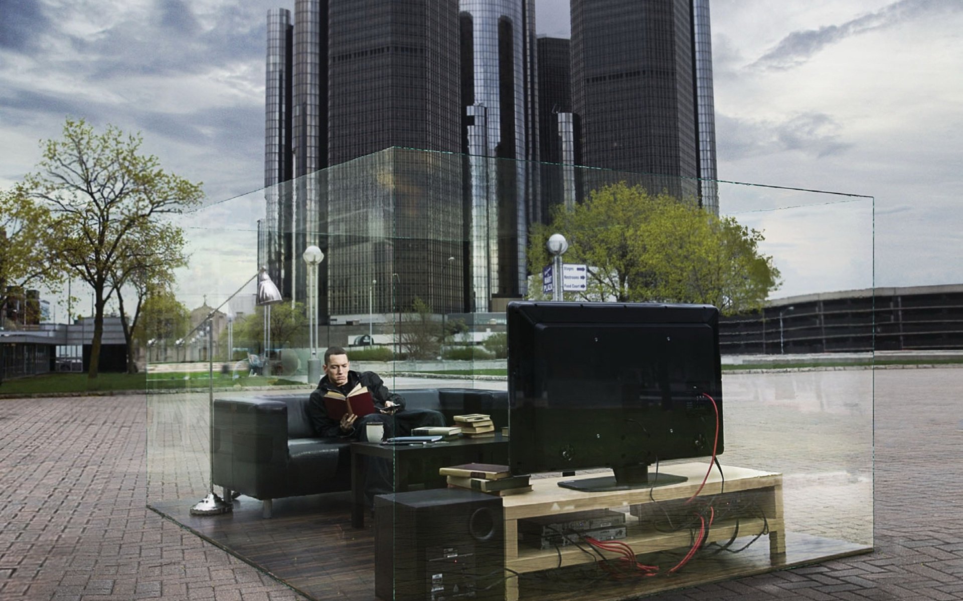 A modern urban setting featuring a transparent living space with a person reading on a couch, surrounded by glass, set against sleek skyscrapers. A striking HD wallpaper for Eminem fans.