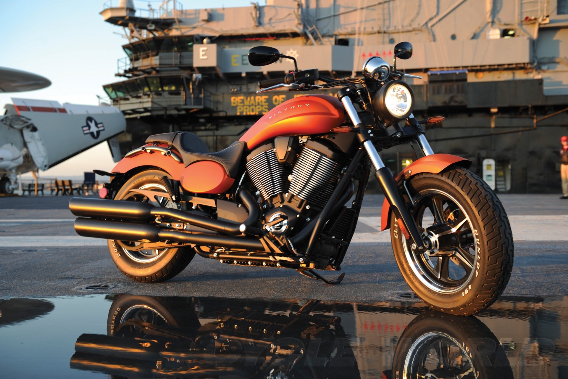 Vehicle — Victory Judge cruiser motorcycle with orange metallic finish reflected on a wet deck in front of an aircraft carrier, HD PC desktop wallpaper background.