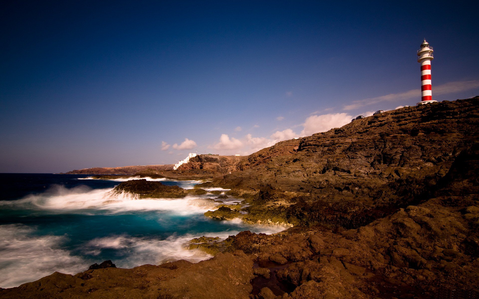A striking lighthouse stands tall against a dramatic seascape, with waves crashing on rocky shores under a clear blue sky. An inspiring man-made structure amidst nature's beauty.