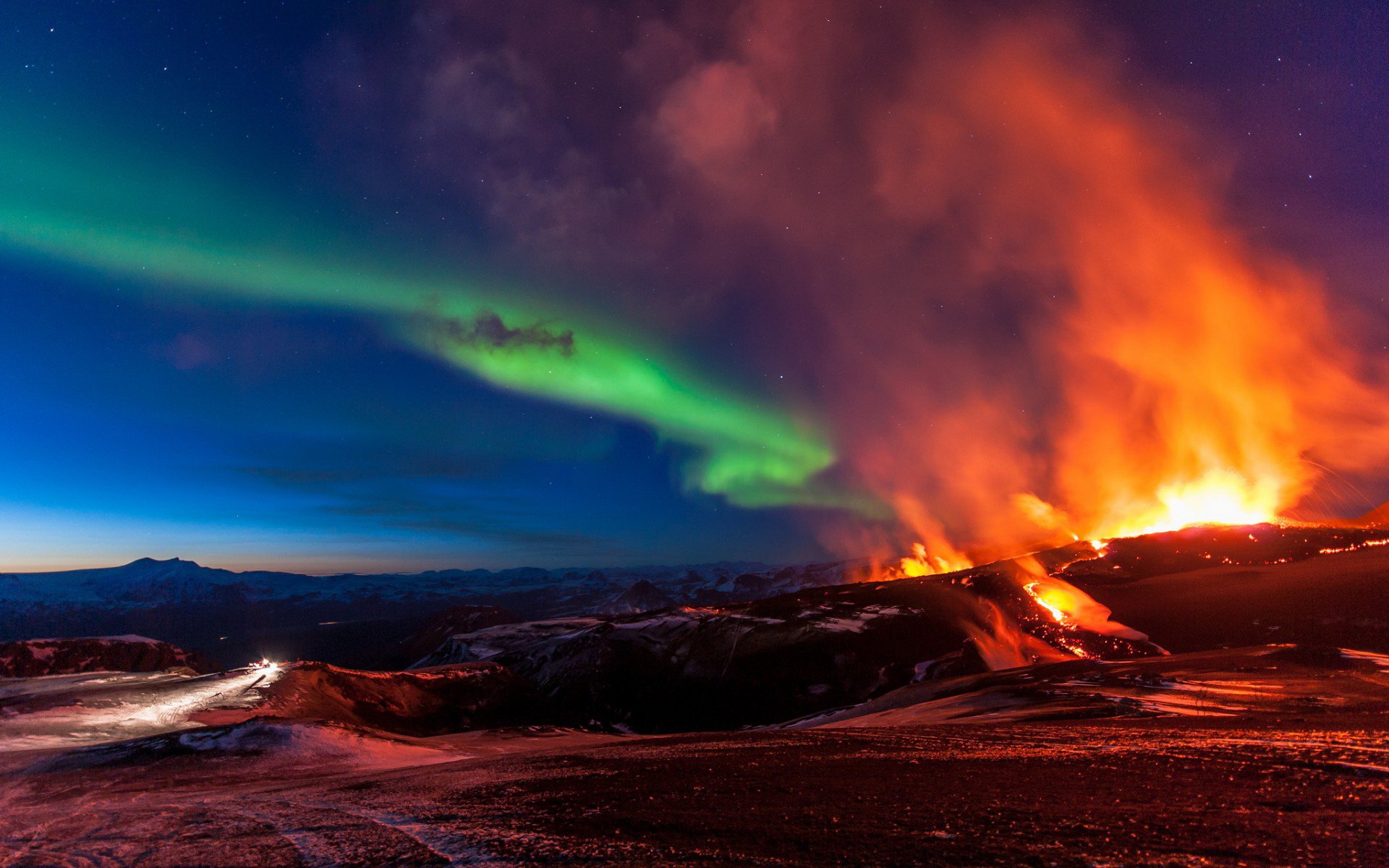 HD desktop wallpaper capturing a vivid aurora borealis glowing above a fiery volcanic eruption in a dramatic natural landscape at night.