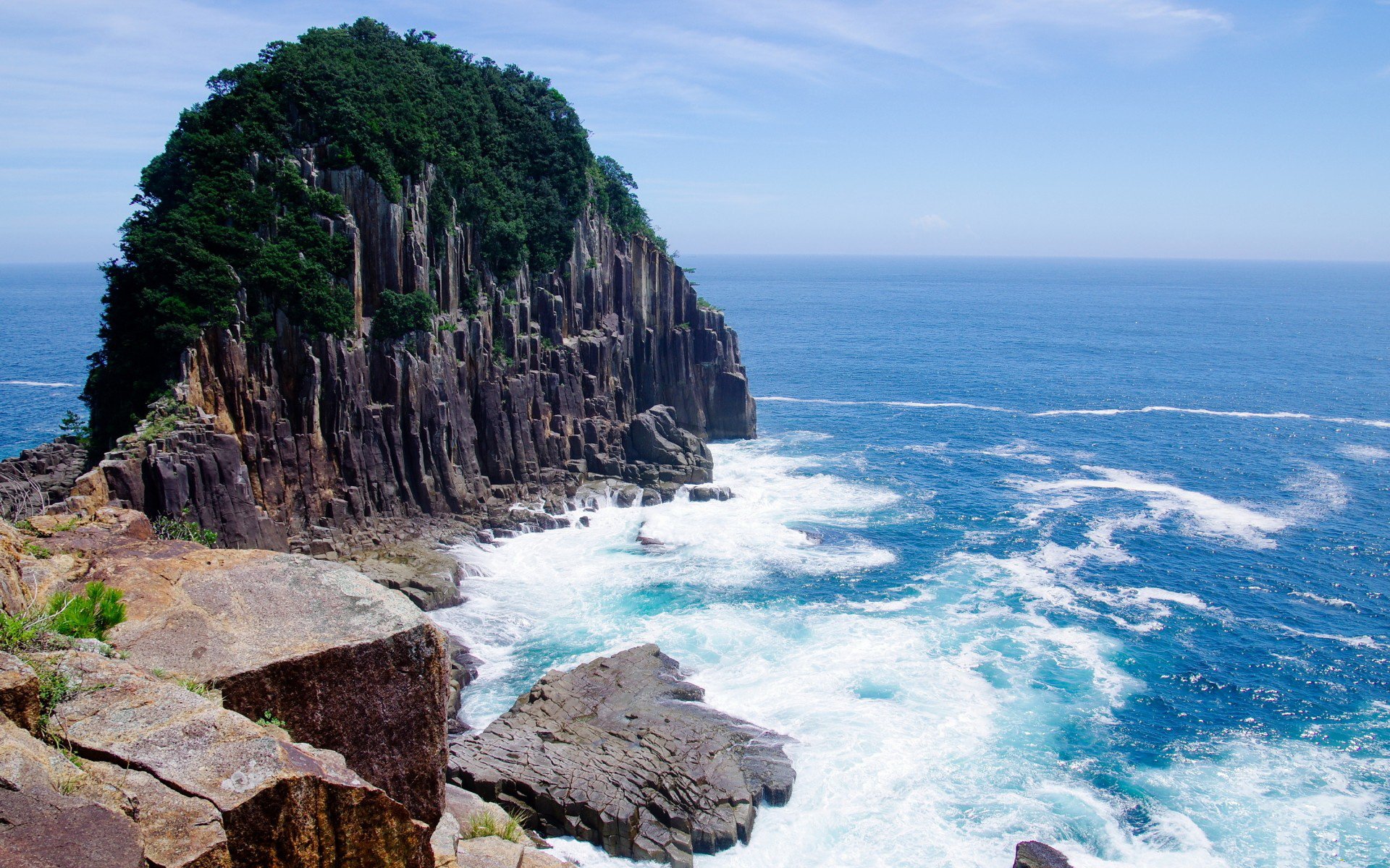 A stunning cliff rises above vibrant blue ocean waves, framed by lush greenery under a clear sky, serving as an inspiring nature-inspired HD desktop wallpaper and background.