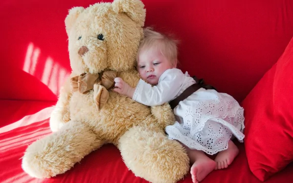 A baby peacefully cuddles a large teddy bear on a vibrant red couch, captured in a heartwarming photograph, creating a charming HD desktop wallpaper and background.