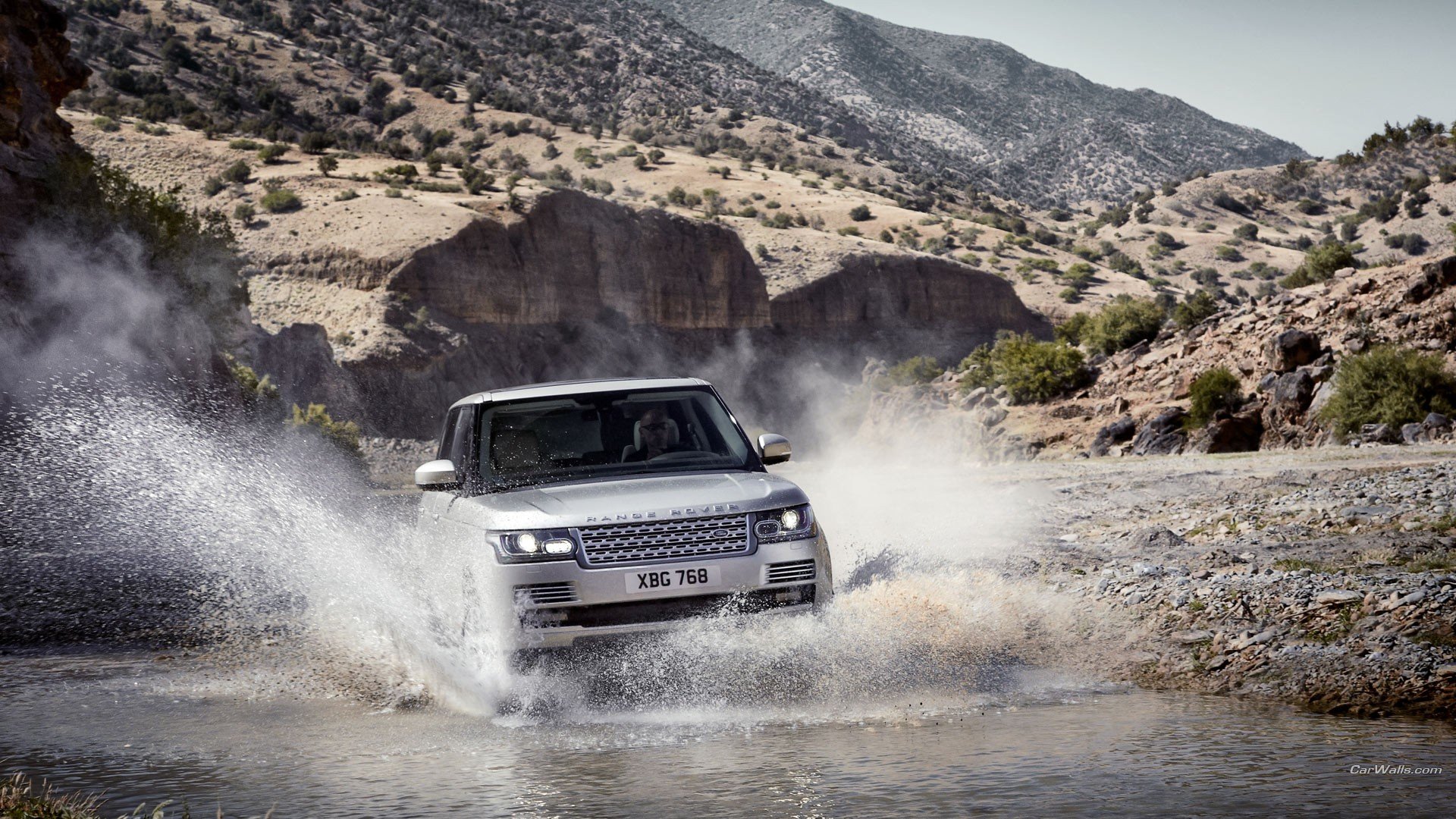 HD desktop wallpaper featuring a Range Rover driving through a rugged, rocky desert landscape with splashing water and mountainous backdrop.