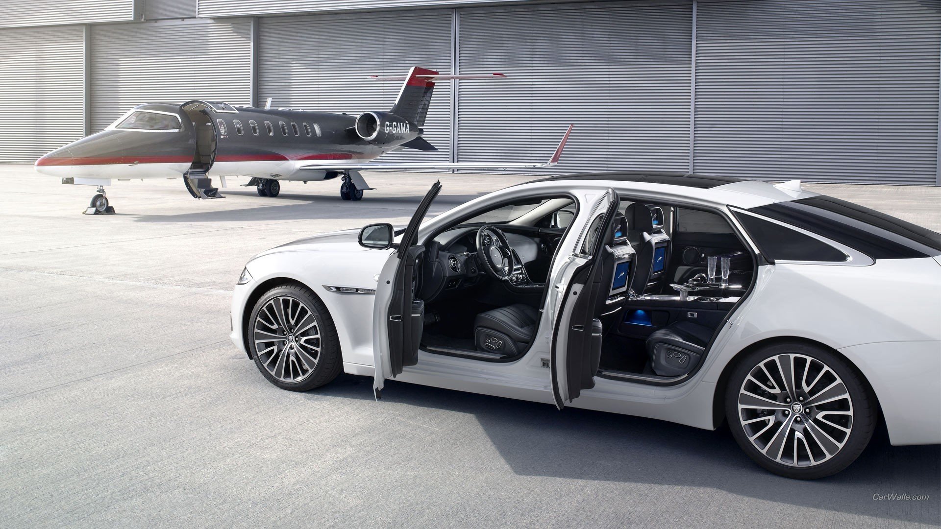 HD PC desktop wallpaper featuring a sleek white Jaguar XJ Ultimate with open doors in a modern hangar alongside a private jet.