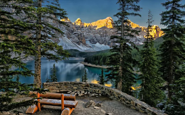 A stunning view of Moraine Lake in Canada, surrounded by towering mountains and dense forests, features a rustic bench overlooking the tranquil blue waters and breathtaking scenery.