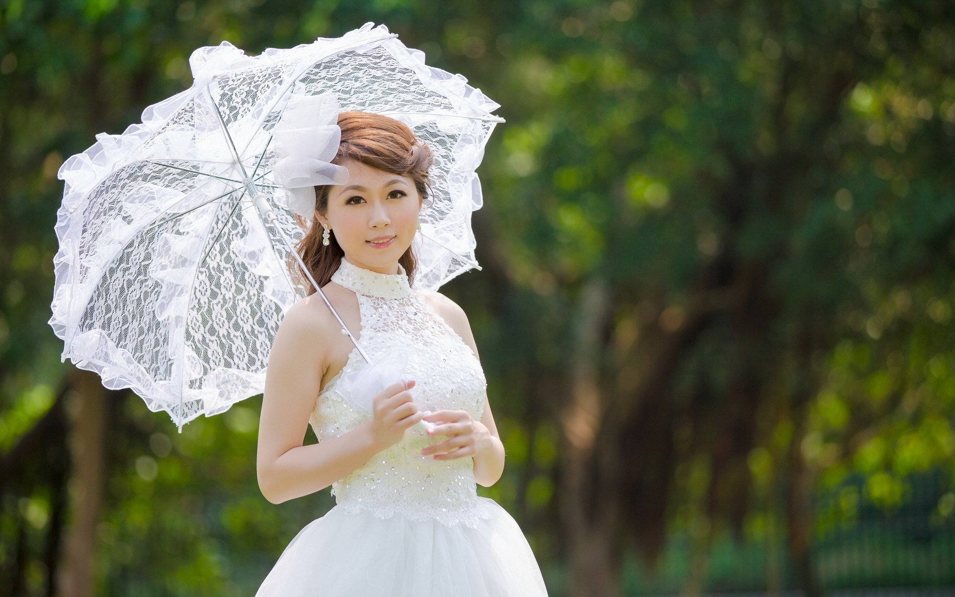 A bride in a stunning white gown holds a lace parasol, standing gracefully amidst a lush green backdrop, creating an enchanting atmosphere for a desktop wallpaper.