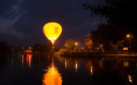 A vibrant yellow hot air balloon is illuminated against a twilight sky, reflecting beautifully on the calm water below, with colorful balloons in the background, creating a serene atmosphere.