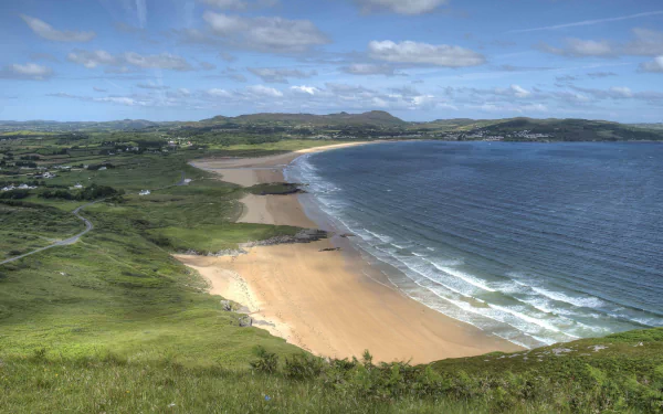 HD PC wallpaper showing a serene coastline with sandy beach, rolling green hills, and gentle ocean waves under a partly cloudy sky.