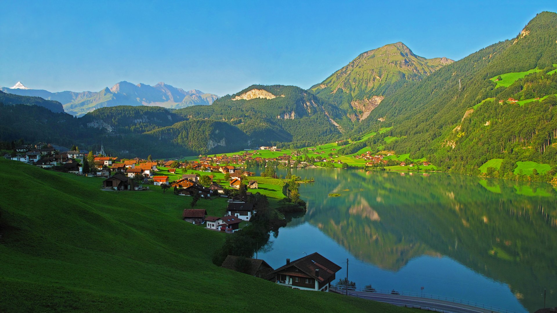 HD desktop wallpaper depicting a serene landscape with a vibrant village nestled between lush green hills and a reflective lake, under a clear blue sky.