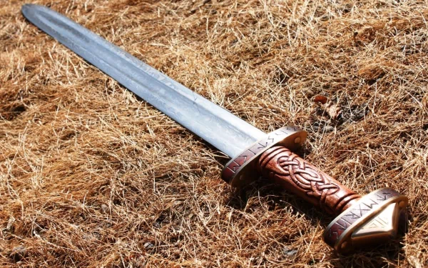HD desktop wallpaper featuring a man-made sword with intricate carvings on the hilt, resting on dry grass.