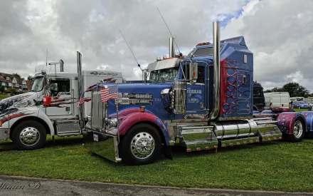A striking Kenworth truck with vibrant colors and decorative details parked on grass, showcasing its impressive design beside another vehicle under a partly cloudy sky.