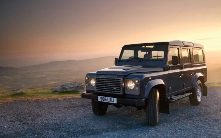 HD PC desktop wallpaper featuring a Land Rover Defender parked on gravel with a scenic sunset landscape in the background.