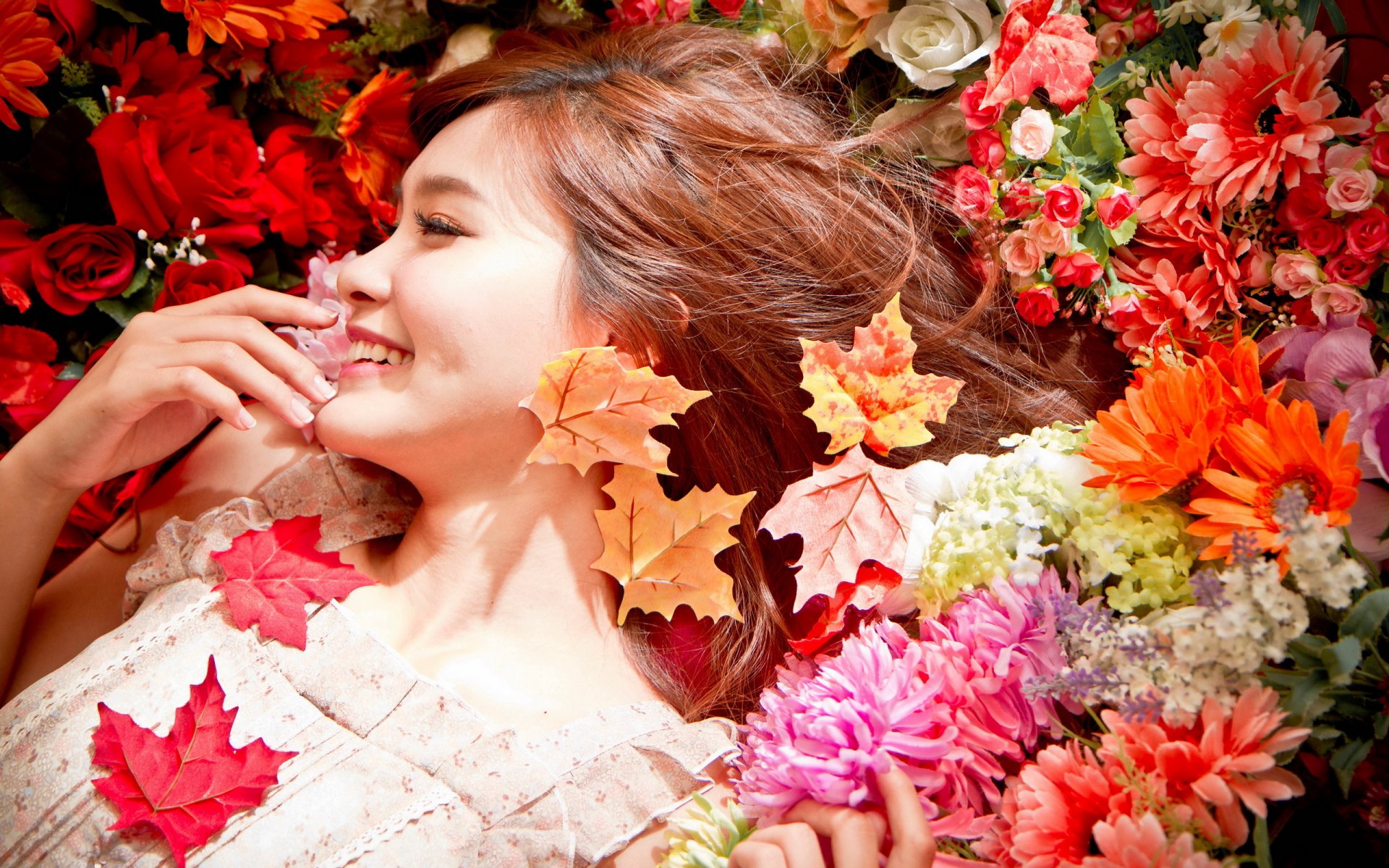 A woman smiles warmly among vibrant flowers and autumn leaves, creating a joyful and serene mood in this HD desktop wallpaper.