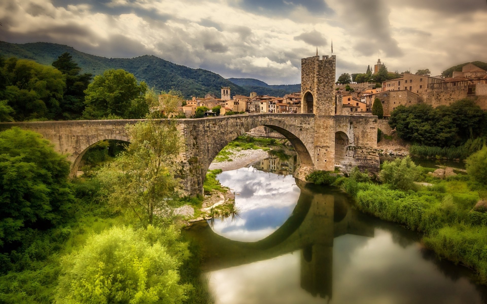 A scenic HD desktop wallpaper featuring a historic stone bridge arching over a tranquil river, surrounded by lush greenery and picturesque mountains in the background.