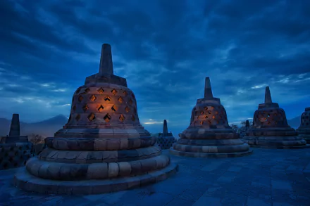 A serene view of Borobudur at dusk, displaying ornate stupas illuminated under a dramatic sky, capturing the rich religious heritage of this iconic site. HD desktop wallpaper and background.
