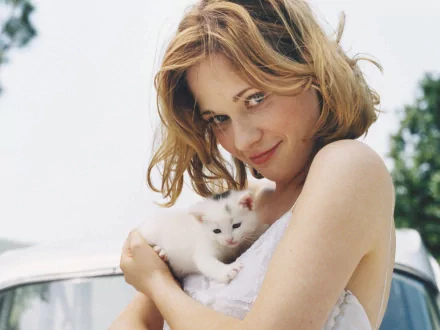 Zooey Deschanel smiles while holding a small white kitten, radiating warmth in a soft, sunlit outdoor setting. A charming HD wallpaper featuring feline companionship.