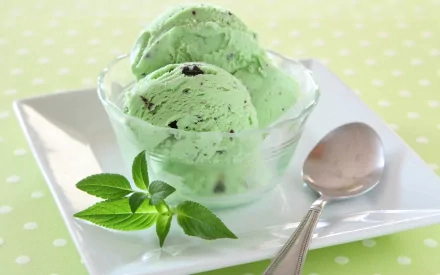 HD desktop wallpaper of mint ice cream in a clear bowl with fresh mint leaves and a spoon, set on a green polka-dotted background.
