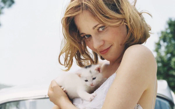 Zooey Deschanel smiles while holding a small white kitten, radiating warmth in a soft, sunlit outdoor setting. A charming HD wallpaper featuring feline companionship.