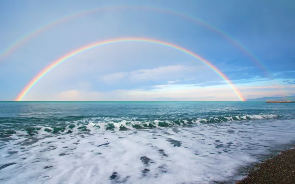 A stunning HD wallpaper of nature featuring a vibrant double rainbow arching over the ocean, with gentle waves lapping the shore under a serene sky.