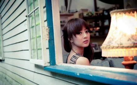 HD PC desktop wallpaper: a woman glances over her shoulder from a blue window frame into a cozy room lit by a vintage table lamp.