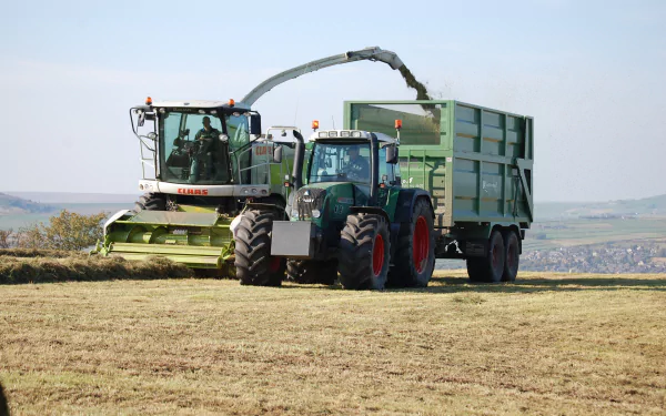 HD desktop wallpaper showing a Fendt tractor towing a large green trailer alongside harvesting machinery in a rural field.