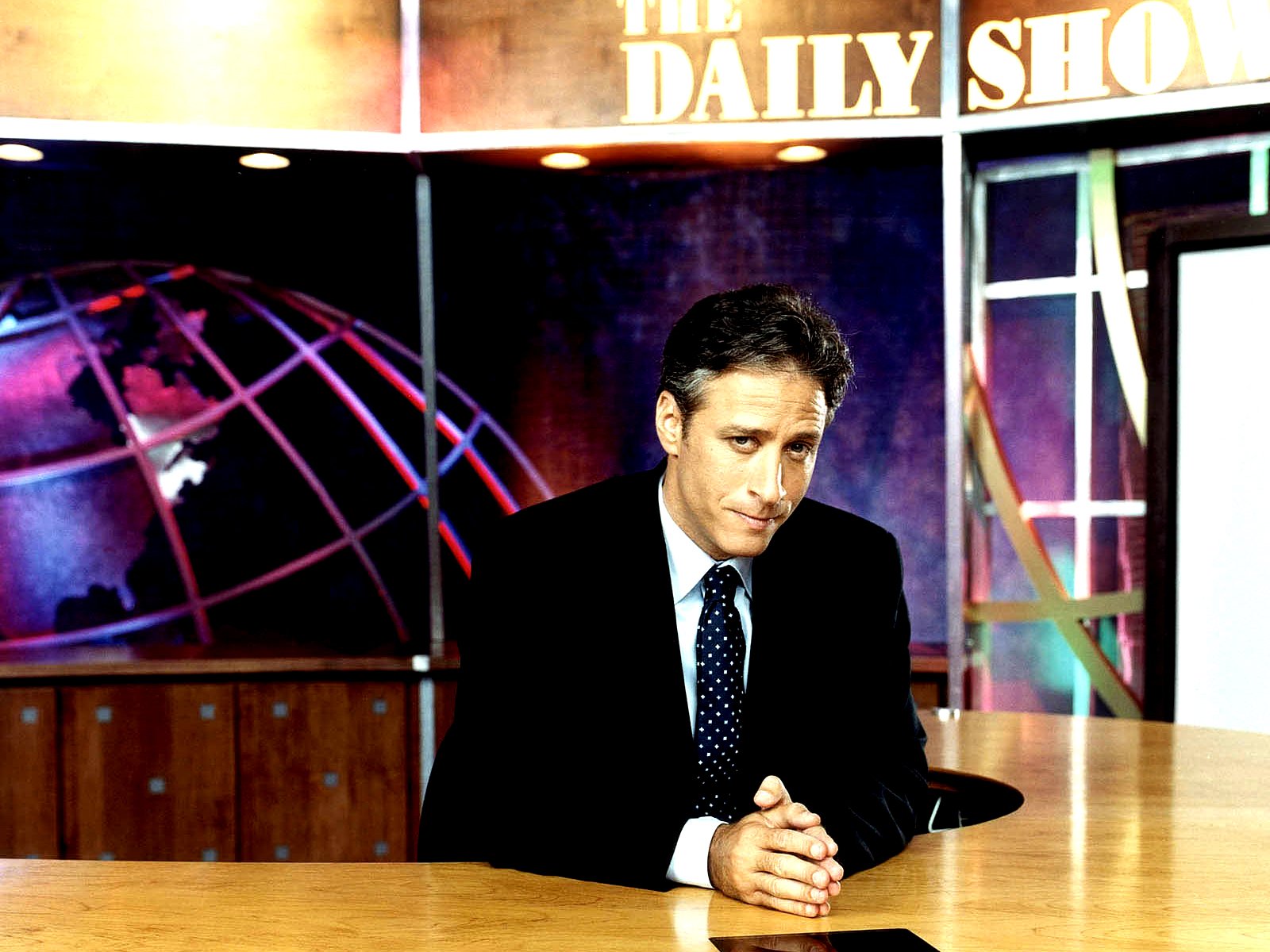 HD desktop wallpaper featuring Jon Stewart on the set of The Daily Show, presenting a thoughtful expression against a vibrant background with a globe motif.