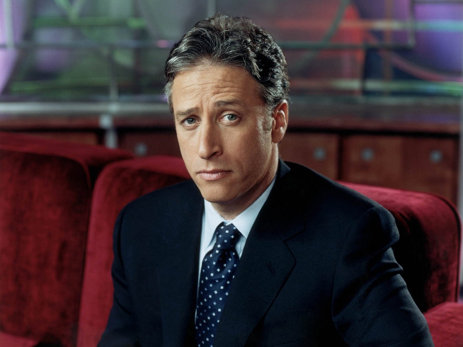 HD wallpaper featuring the host from The Daily Show with Jon Stewart in a suit, poised against a plush red backdrop.