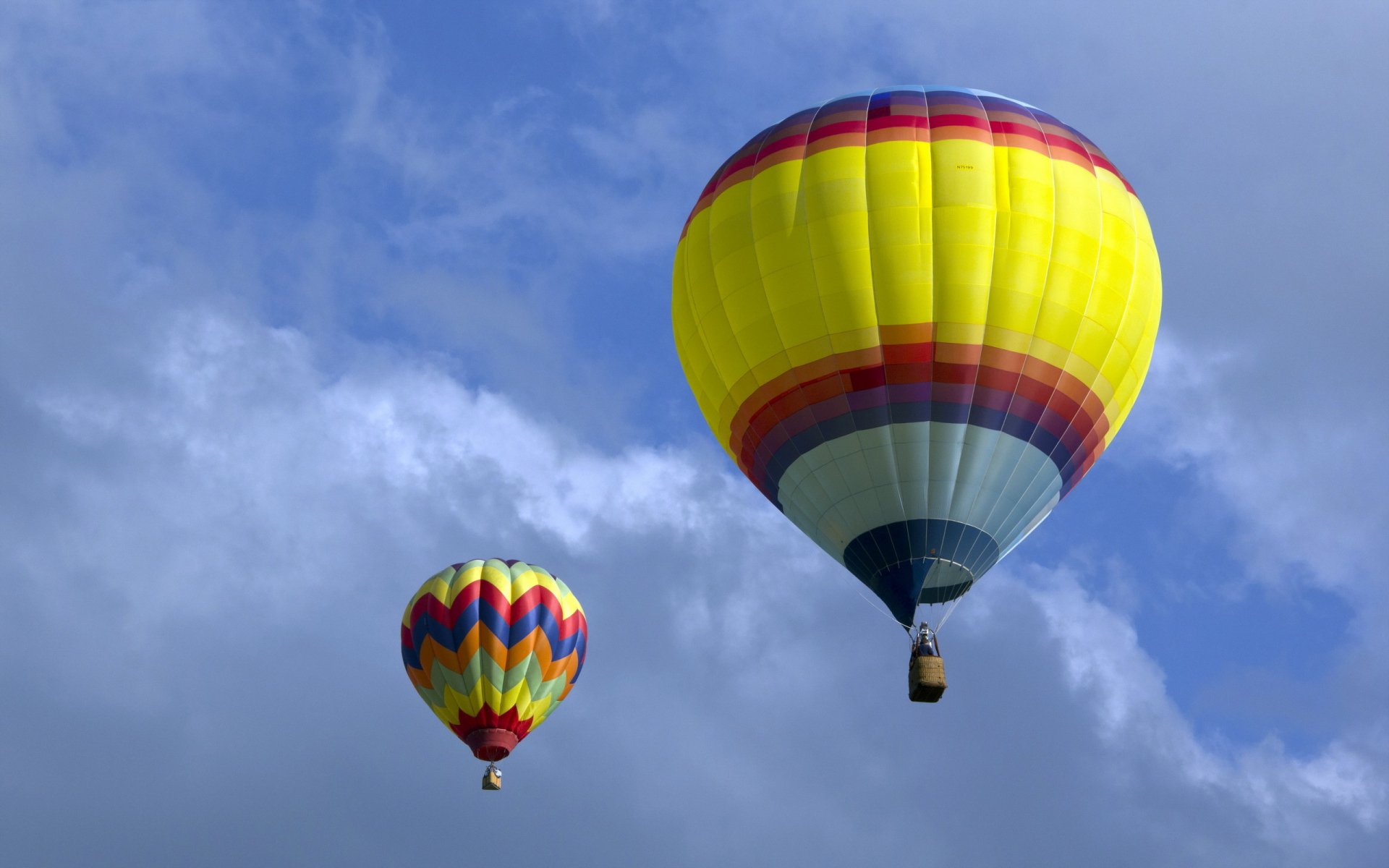 HD desktop wallpaper featuring two colorful hot air balloons floating against a bright blue sky with scattered clouds.