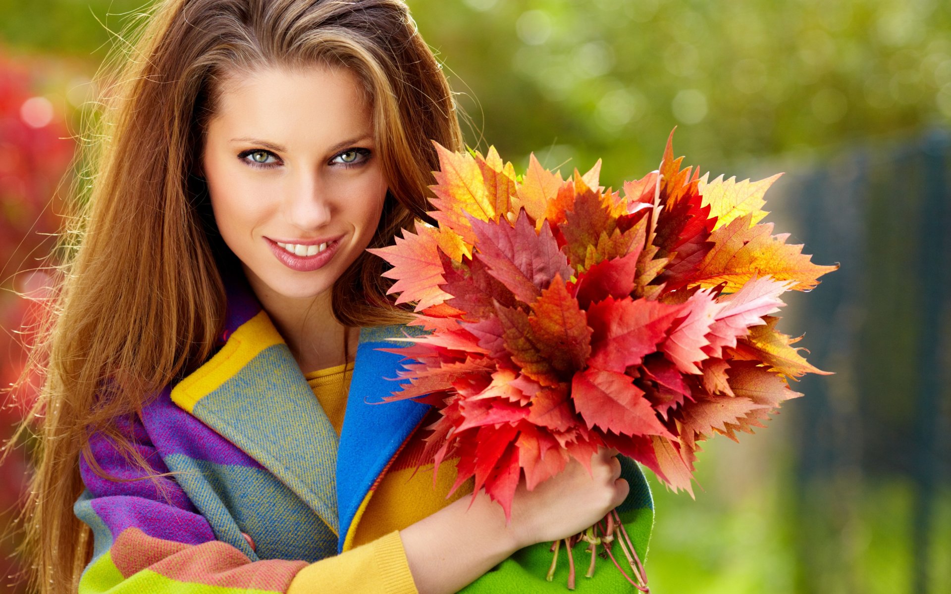 A woman named Izabela Margiera smiles while holding a vibrant bouquet of autumn leaves, dressed in a colorful coat against a natural background. HD desktop wallpaper and background.