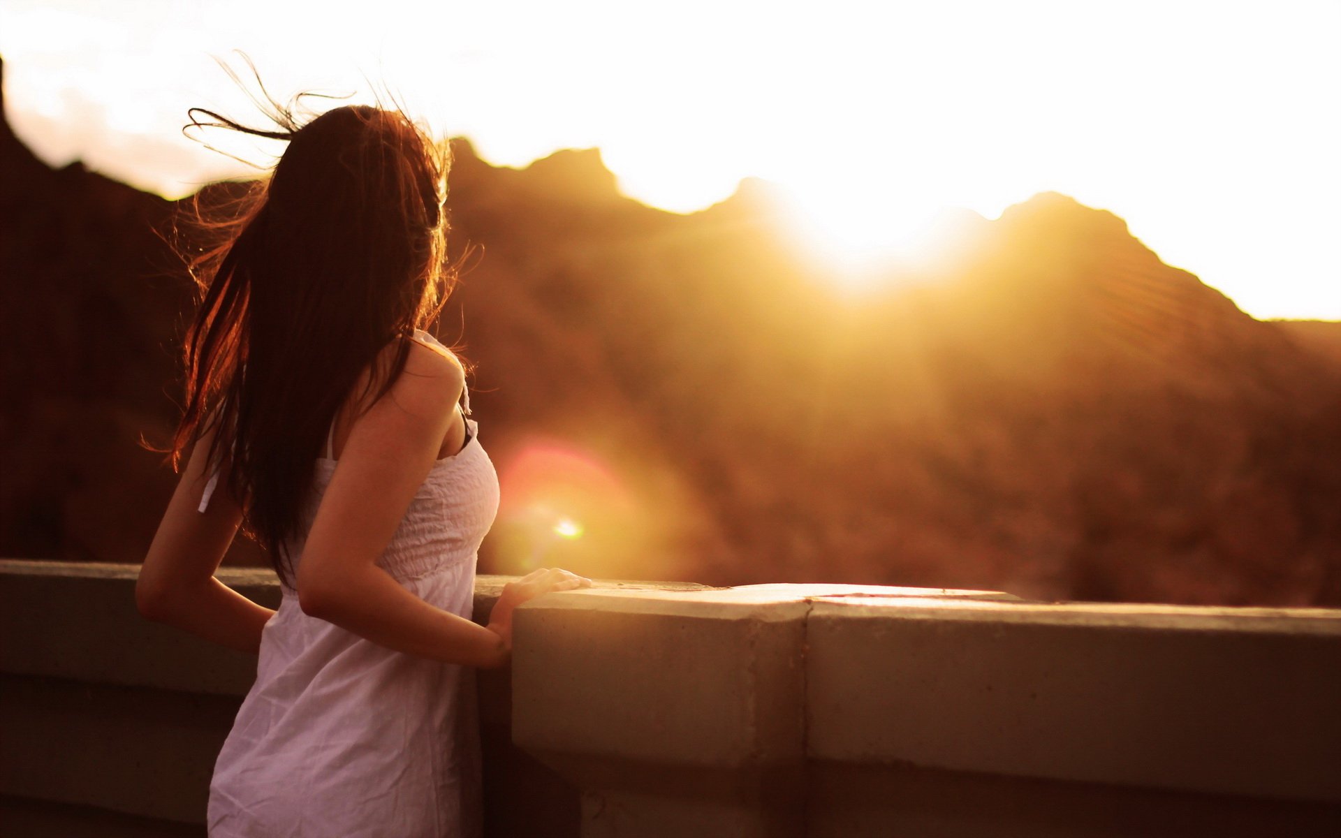 A woman stands on a balcony, gazing at a stunning sunset over mountains, her hair gently blowing in the breeze. This HD wallpaper captures a serene mood and breathtaking scenery.