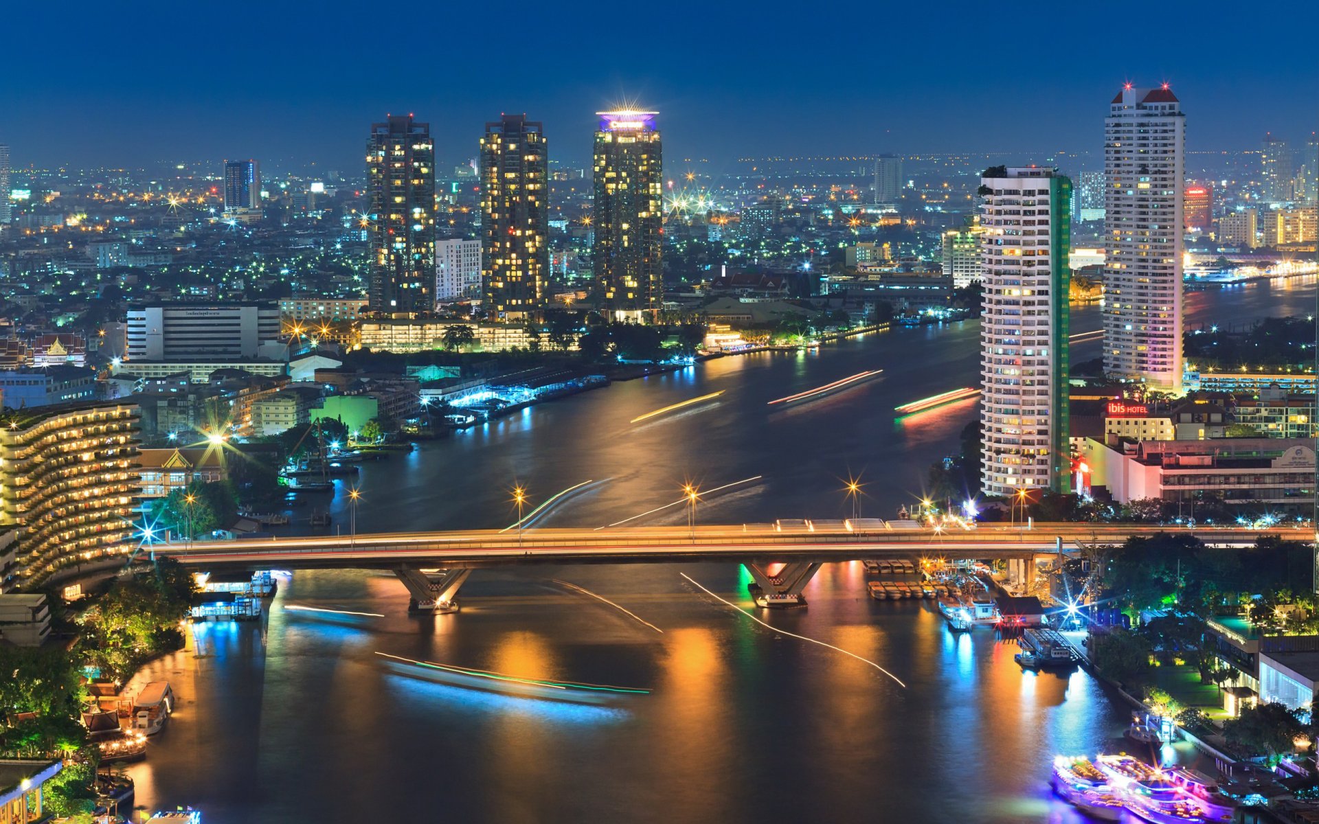 Nighttime view of Bangkok’s illuminated skyline with high-rise buildings and bridges over a river, captured in HD for a vibrant desktop wallpaper background.