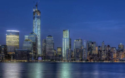 HD desktop wallpaper of New York's Manhattan skyline at dusk, a man-made panorama with One World Trade Center towering above the waterfront, lights reflecting on the water.