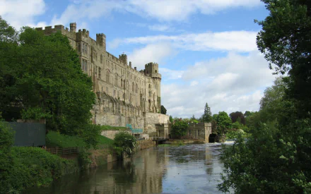 HD PC desktop wallpaper showing the man-made Warwick Castle standing beside a calm river, surrounded by lush greenery under a partly cloudy sky.