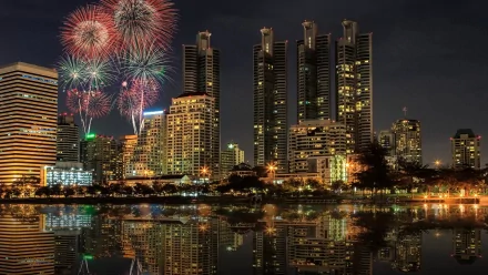 A vibrant night scene of Bangkok's skyline, featuring modern skyscrapers illuminated by city lights, with colorful fireworks bursting above and reflections shimmering on the water.