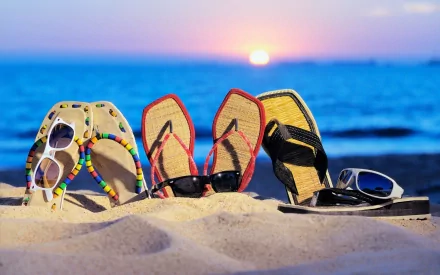 Three colorful thongs (flip‑flops) and sunglasses planted in sand at sunset on a beach — humorous summer holiday photography, 2K Quad HD PC desktop wallpaper/background.