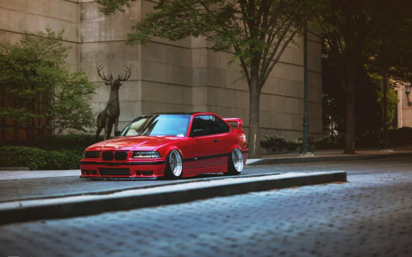 A striking red BMW parked on a city street, framed by trees and a nearby statue, enhances this HD desktop wallpaper and background.